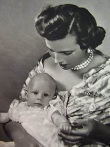 Prinses Lilian in gebloemde jurk en met parelketting en parel uit de tiaraals broche, met dochter prinses Maria-Esmeralda op schoot
