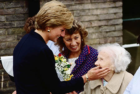 Een glimlachende prinses Diana in zwart jasje aait de wang van een oudere mevrouw tijdens een werkbezoek